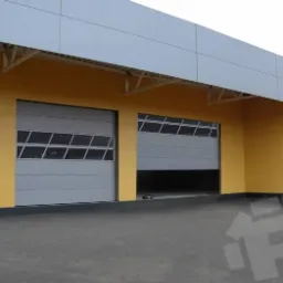 Szara brama przemysłowa z oknami częściowo otwarta w żółto-szarym budynku przemysłowym, widoczny fragment wnętrza.