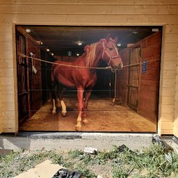 Mariusz Soszyński Przedsiębiorstwo Handlowo Usługowe - Nowa brama garażowa z fotorealistycznym nadrukiem przedstawiającym konia w stajni, zamontowana w drewnianym garażu z widocznymi elementami montażowymi u dołu.