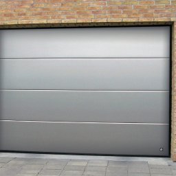 Mariusz Soszyński Przedsiębiorstwo Handlowo Usługowe - Zamknięta, segmentowa brama garażowa w kolorze srebrnym, z czterema poziomymi panelami, zamontowana w murze z cegły klinkierowej, oświetlona przez dwa zewnętrzne kinkiety.