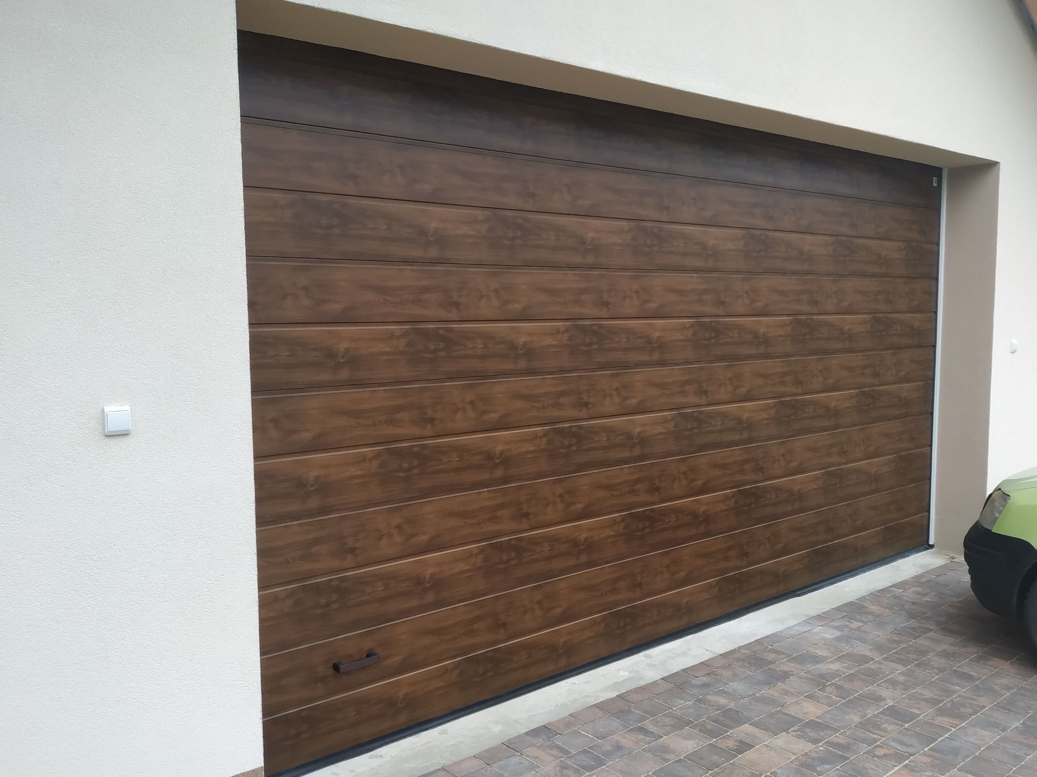 Zamknięta, segmentowa brama garażowa w kolorze ciemnego drewna z poziomymi panelami, zamontowana w jasnym garażu, widoczny fragment kostki brukowej i samochodu.