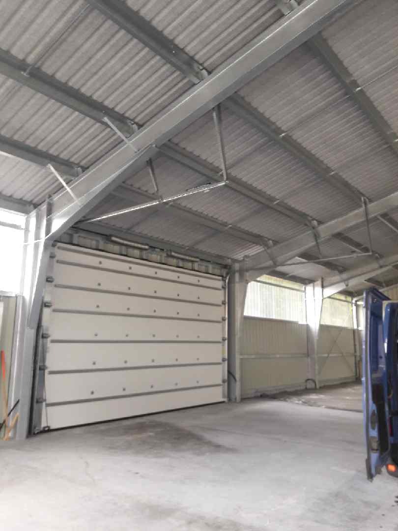 Zamknięta, segmentowa brama przemysłowa wewnątrz hali z metalową konstrukcją dachu, widoczny fragment niebieskiego samochodu dostawczego.