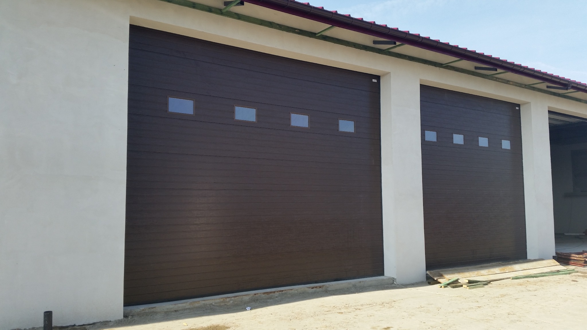 Dwie brązowe segmentowe bramy z czterema małymi kwadratowymi okienkami, zamontowane w jasnym budynku o płaskim dachu z widocznymi elementami konstrukcyjnymi.