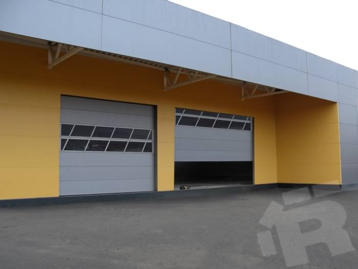 Szara brama przemysłowa z oknami częściowo otwarta w żółto-szarym budynku przemysłowym, widoczny fragment wnętrza.
