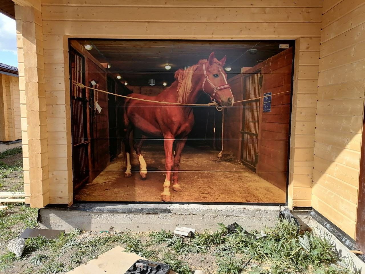 Nowa brama garażowa z fotorealistycznym nadrukiem przedstawiającym konia w stajni, zamontowana w drewnianym garażu z widocznymi elementami montażowymi u dołu.