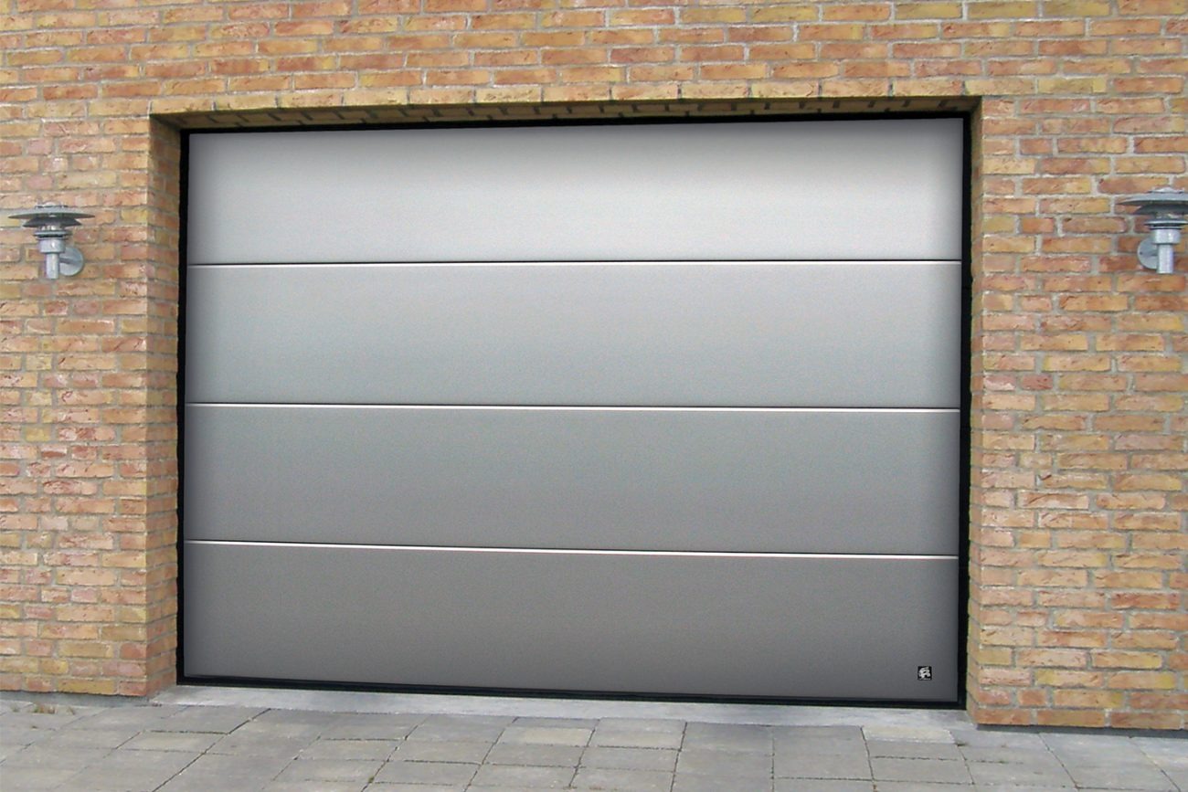 Zamknięta, segmentowa brama garażowa w kolorze srebrnym, z czterema poziomymi panelami, zamontowana w murze z cegły klinkierowej, oświetlona przez dwa zewnętrzne kinkiety.