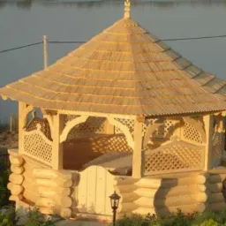 Drewniana altana z dachem krytym strzechą, z ażurowymi ściankami i małą bramką, ustawiona w ogrodzie z widokiem na jezioro.