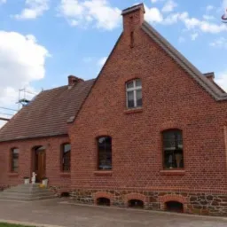 Ceglany dom z czerwoną dachówką, widoczne rusztowania z boku, kamienna podmurówka, logo firmy w prawym górnym rogu.