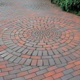 Ułożona z czerwono-szarych cegieł nawierzchnia z dekoracyjnym wzorem koła i rozet, obrzeża z zielenią.