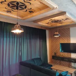 Drewniane kasetony sufitowe z ciemnym wzorem roślinnym, oświetlone wiszącymi lampami w salonie z zielonymi zasłonami i narożnikiem.