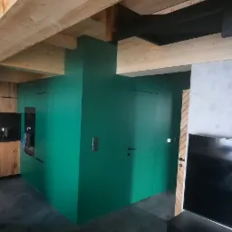 Nowoczesne wnętrze: kuchnia z drewnianymi szafkami, intensywnie zielona zabudowa z ukrytymi drzwiami, betonowe schody z czarnymi stopniami i drewniane belki stropowe.