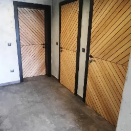 Trzy nowoczesne drzwi wewnętrzne z drewnianymi panelami ułożonymi w jodełkę, czarne ramy i klamki, szare ściany i podłoga, drewniany sufit z oświetleniem punktowym.