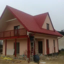 Montaż rynny przy nowo wybudowanym domu z czerwonym dachem i balkonem; w tle widoczny kościół z dwiema wieżami.