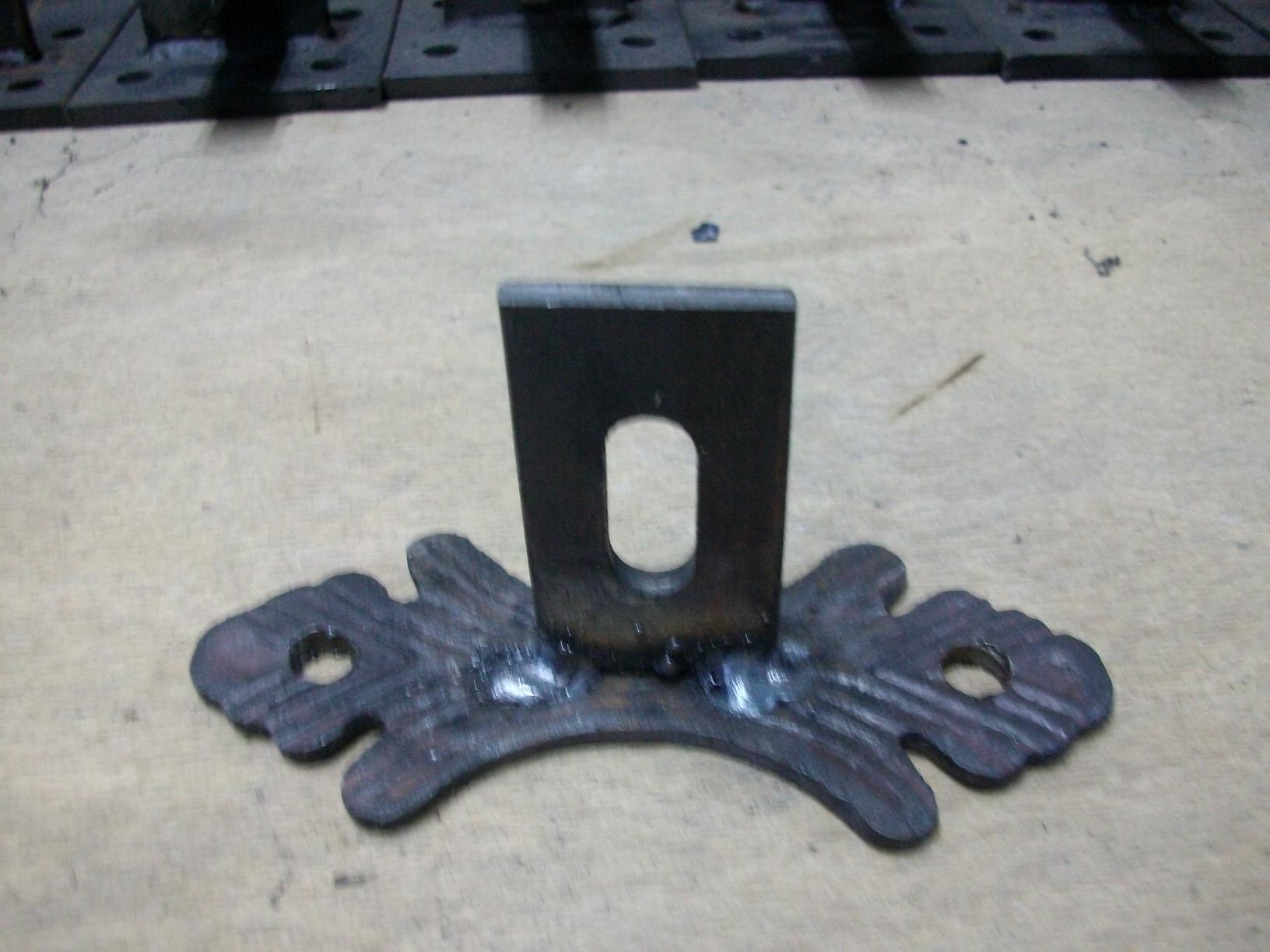 Surowy, metalowy uchwyt montażowy przęseł z widocznymi spawami i otworem, leżący na drewnianej powierzchni roboczej, z innymi uchwytami w tle.