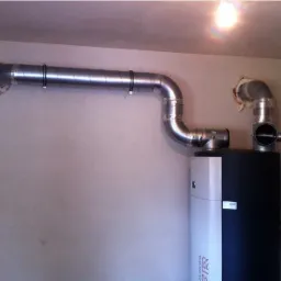 Czarno-biała pompa ciepła z podłączonymi srebrnymi rurami wentylacyjnymi, zamontowana przy białej ścianie z widocznymi śladami montażu.