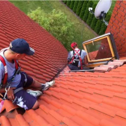 Dwóch monterów na dachu w Mławie instaluje okno dachowe, zabezpieczeni uprzężami. Widoczny fragment dachu z czerwonej dachówki i satelita w tle.