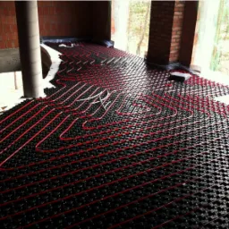 Montaż ogrzewania podłogowego REHAU z czerwonymi rurami na czarnej płycie systemowej w surowym stanie budynku z widocznymi ceglanymi filarami i drzewami w tle.