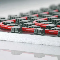 Fragment systemu ogrzewania podłogowego REHAU z czerwonymi rurkami umieszczonymi w czarnych uchwytach na białym styropianie, widoczne oznaczenia na rurkach.