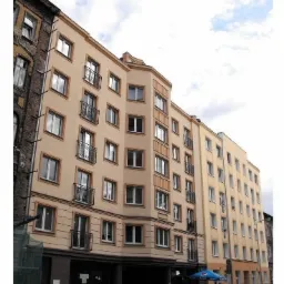 Wielopiętrowy budynek mieszkalny po renowacji elewacji, z widoczną różnicą w kolorystyce między odnowioną częścią a sąsiednim budynkiem, z balkonami z metalowymi barierkami i plastikowymi oknami.