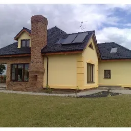 Żółty dom jednorodzinny z ciemną dachówką, ceglanym kominem i kolumnami wejściowymi, panele słoneczne na dachu, widok z trawnika.