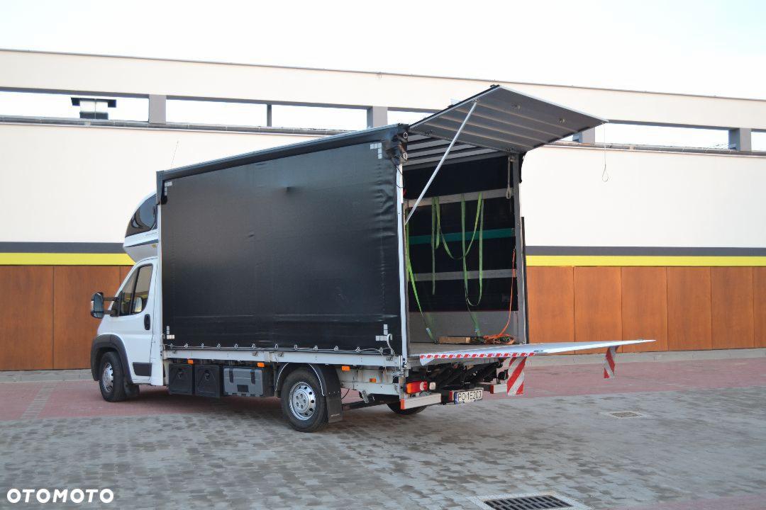 Biały samochód dostawczy z otwartą czarną plandeką, widoczne pasy mocujące ładunek wewnątrz, tło stanowi biało-brązowy budynek.