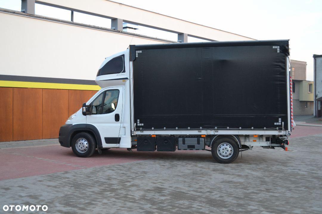 Biały samochód dostawczy z czarną plandeką na tle budynku z drewnianą elewacją.