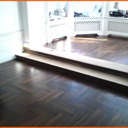 Ciemna, polerowana podłoga parkietowa ułożona we wzór jodełki francuskiej, z widocznym progiem i fragmentem pianina w tle, oświetlona naturalnym światłem wpadającym przez okno.