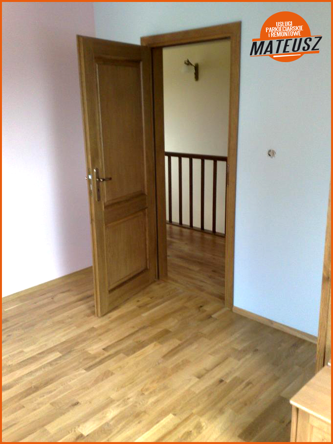 Otwarta drewniana dębowa ościeżnica z zamontowanymi drzwiami, widok na świeżo wycyklinowany parkiet i klatkę schodową w tle.