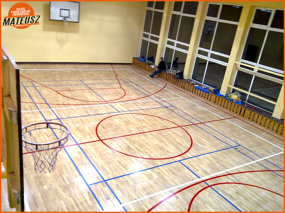 Widok z góry na odnowioną drewnianą podłogę hali sportowej z wyraźnie odmalowanymi liniami boiska do koszykówki, z koszem po lewej stronie i oknami w tle, gdzie widać siedzącą osobę.