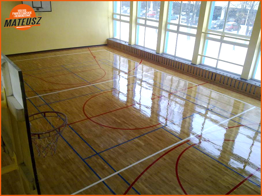 Błyszcząca, odnowiona drewniana podłoga sali gimnastycznej z liniami boiska do koszykówki, widok z góry na kosz i okna z zimowym krajobrazem za nimi.