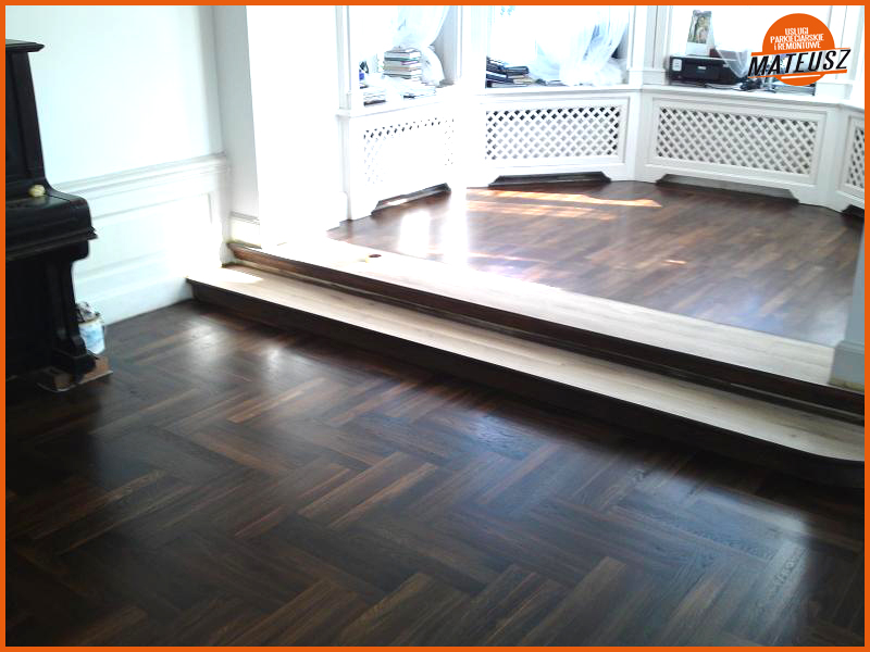 Ciemna, polerowana podłoga parkietowa ułożona we wzór jodełki francuskiej, z widocznym progiem i fragmentem pianina w tle, oświetlona naturalnym światłem wpadającym przez okno.