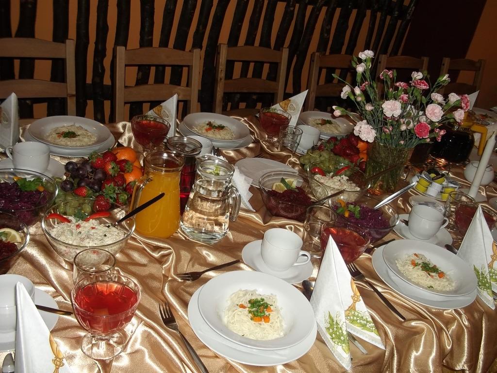 Uroczysty stół nakryty złotym obrusem, zastawiony talerzami z daniami, szklankami z napojami, miskami z sałatkami, owocami i wazonem z kwiatami, przygotowany do obsługi imprezy.