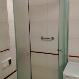 Uslugi glazurnicze - Wykończona łazienka z kabiną prysznicową z półprzezroczystymi szybami i chromowanymi okuciami, widoczna umywalka i pralka. Białe płytki z bordową fugą.