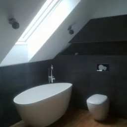 Nowoczesna łazienka na poddaszu z wolnostojącą wanną, drewnopodobną podłogą i oknem dachowym. Ciemne płytki na ścianach.