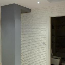 Uslugi glazurnicze - Wykończone wnętrze z dekoracyjną białą cegłą na ścianie, szarą kolumną i widocznym wejściem do łazienki. Na podłodze widoczne materiały budowlane.