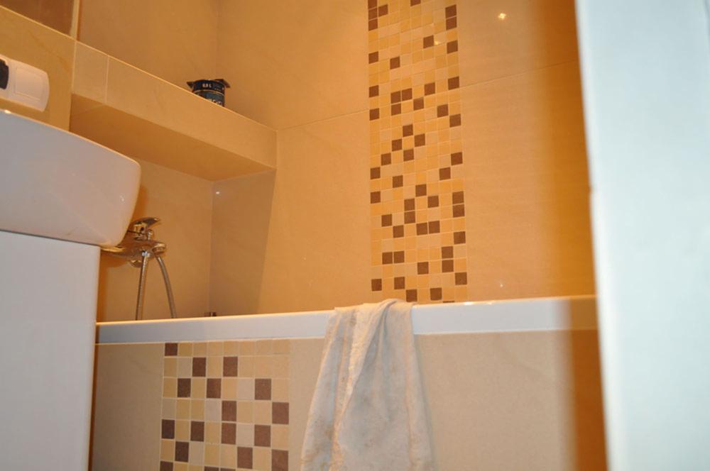 Beżowa łazienka z mozaiką w odcieniach brązu i beżu. Widoczna wanna z ręcznikiem, umywalka i armatura. Na ścianie półka z kosmetykami.