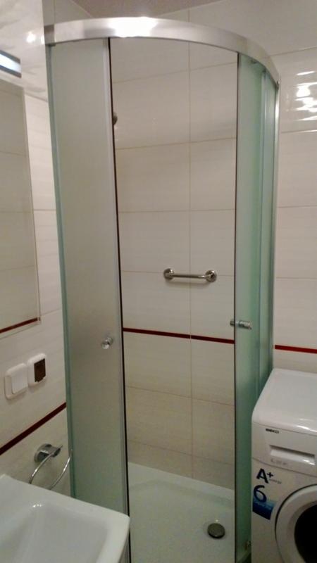 Wykończona łazienka z kabiną prysznicową z półprzezroczystymi szybami i chromowanymi okuciami, widoczna umywalka i pralka. Białe płytki z bordową fugą.