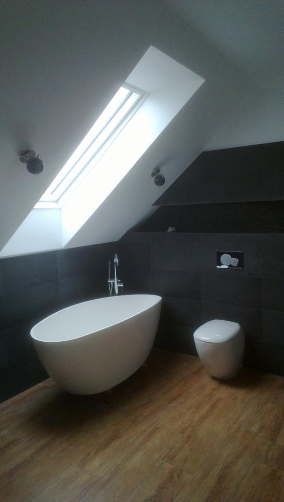 Nowoczesna łazienka na poddaszu z wolnostojącą wanną, drewnopodobną podłogą i oknem dachowym. Ciemne płytki na ścianach.
