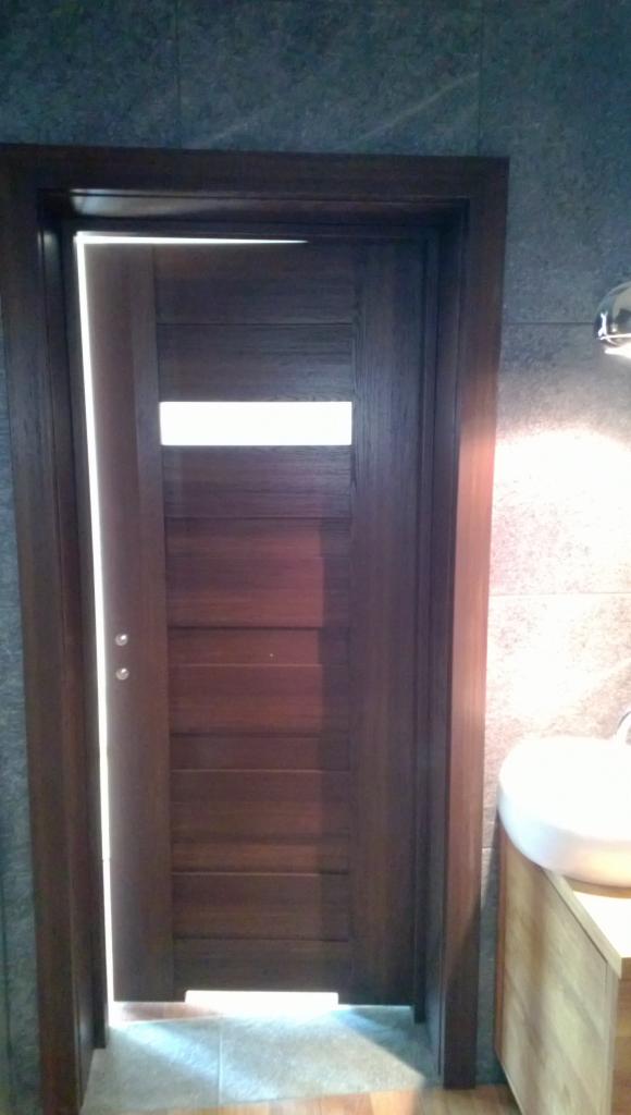 Nowoczesne, ciemne drewniane drzwi z poziomymi panelami i przeszkleniem, w łazience z szarą płytką i białą umywalką na drewnianej szafce.