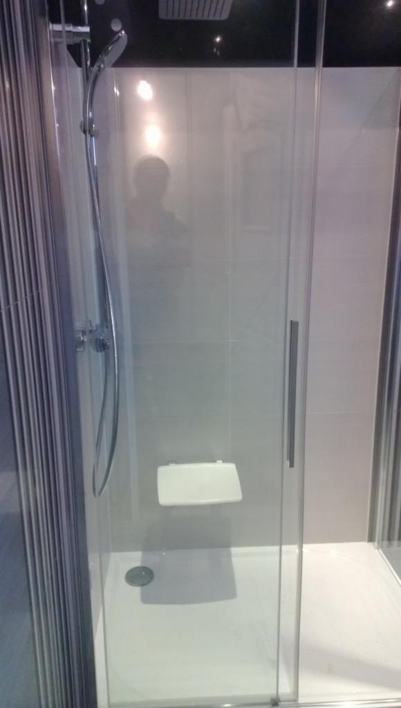 Nowoczesna kabina prysznicowa z siedziskiem, chromowaną armaturą i przesuwnymi drzwiami, wykończona jasnymi płytkami. Minimalistyczny design.