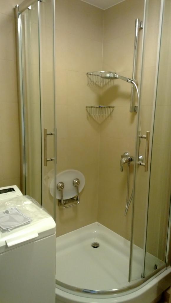 Nowoczesna kabina prysznicowa z siedziskiem i półkami w jasnej łazience. Widoczna pralka z dokumentacją. Minimalistyczny design, praktyczne rozwiązania.