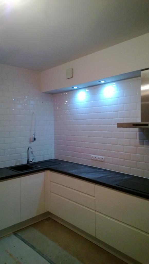 Wykończona kuchnia z białymi, błyszczącymi płytkami typu metro na ścianie, czarnym blatem i jasnymi szafkami. Widoczne oświetlenie podszafkowe i fragment okapu.