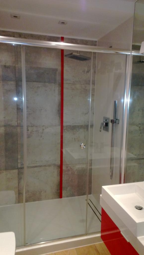 Nowoczesna kabina prysznicowa z przesuwnymi drzwiami i czerwonym akcentem, widoczna umywalka z szafką w kolorze czerwonym, szare płytki na ścianie.