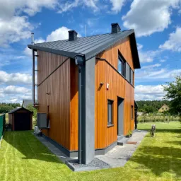 Nowoczesny dom z elewacją z pionowych paneli drewnianych, szare elementy wykończeniowe, ciemna dachówka, widok na zadbany trawnik i błękitne niebo z chmurami.