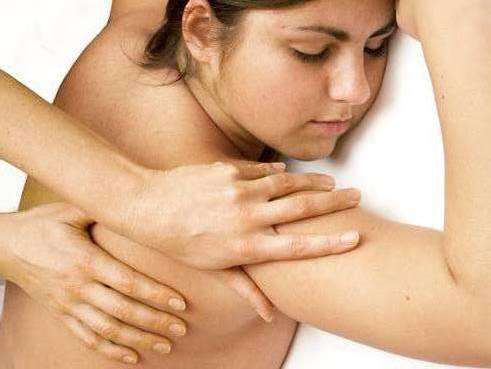 Kobieta otrzymuje masaż ramion i pleców, z zamkniętymi oczami i rozluźnioną twarzą, dłonie masażysty delikatnie ugniatają mięśnie.