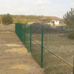 Zielone ogrodzenie panelowe z widocznymi słupkami na nierównym, piaszczystym terenie, z domem w tle i fragmentem lasu.