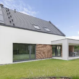 Nowoczesny dom jednorodzinny z grafitową dachówką, białym tynkiem i elementami z cegły klinkierowej, zadaszony taras z meblami ogrodowymi oraz zadbany trawnik.