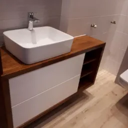 Nowoczesna łazienka: biała, kwadratowa umywalka nablatowa z chromowaną baterią na drewnianym blacie z białymi szufladami, widoczne fragmenty toalety i płytek.