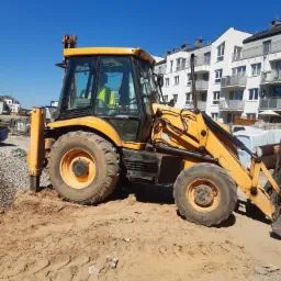 Żółta koparko-ładowarka JCB na piaszczystym terenie budowy z nowym budynkiem mieszkalnym w tle, widok z boku w słoneczny dzień.