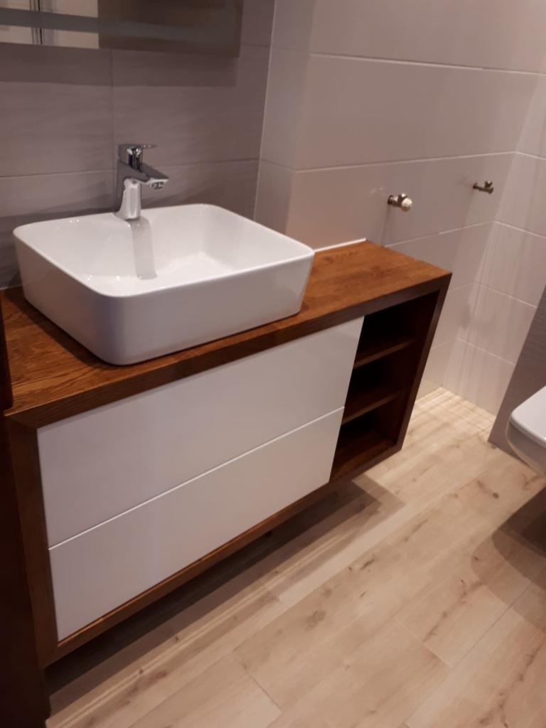 Nowoczesna łazienka: biała, kwadratowa umywalka nablatowa z chromowaną baterią na drewnianym blacie z białymi szufladami, widoczne fragmenty toalety i płytek.