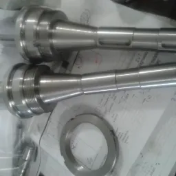 Dwa metalowe trzpienie tokarskie o różnych długościach leżące na arkuszu z technicznym rysunkiem, obok nakrętka z gwintem wewnętrznym.